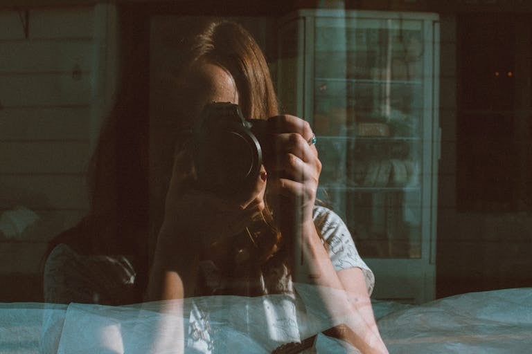 A woman holding a camera captures her reflection through a glass window, creating a reflective portrait.