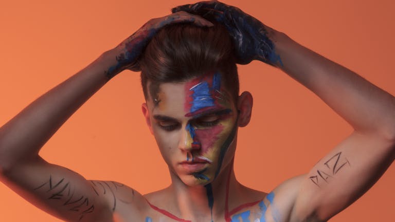 A young man with colorful face paint and an orange background, hands on head.
